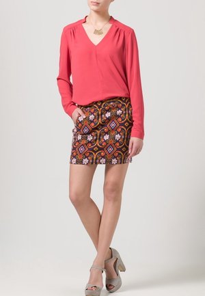 Blouse rouge à manches longues avec un col en V, associée à une mini-jupe à motifs avec des designs géométriques et des accents floraux. Escarpins gris.
