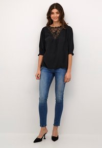 Blusa nera con dettagli in pizzo al collo, maniche a tre quarti e vestibilità ampia, abbinata a jeans skinny blu e tacchi neri.