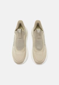 Ζευγάρι μπεζ αθλητικά παπούτσια πλεκτού τύπου με λευκές σόλες, υφή από πλέγμα στην επάνω πλευρά και θηλιά στο πίσω μέρος, απεικονισμένα από πάνω σε λευκό φόντο.