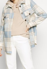 Flanellen jack in crème, blauw en tan geruit patroon. Bevat twee borstzakken, knopen en een capuchon. Draagt over een beige gebreide trui.