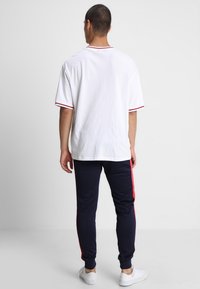 Witte t-shirt met rode afwerking, gecombineerd met marineblauwe sweatpants met rode zijstrepen. Schoeisel is witte sneakers.