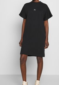 Personne portant une robe noire à manches courtes avec un petit texte blanc "WON HUNDRED" sur la poitrine et des bottines beiges, se tenant sur un sol gris.