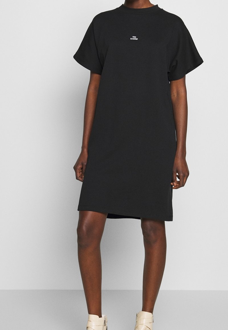 Personne portant une robe noire à manches courtes avec un petit texte blanc "WON HUNDRED" sur la poitrine et des bottines beiges, se tenant sur un sol gris.