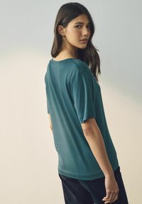T-shirt à manches courtes turquoise en tissu doux, avec un col rond et une coupe décontractée, associé à un pantalon sombre sur un fond neutre.