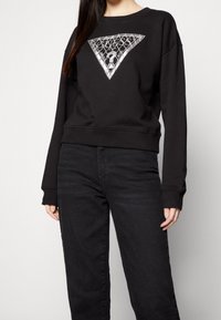 Sudadera negra con un gráfico triangular plateado en el frente, con puños y dobladillo acanalados, combinada con vaqueros oscuros.