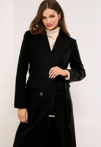 Mujer vestida con un abrigo de lana negro con cinturón sobre un suéter de cuello alto beige, con el cabello suelto castaño y pendientes de aro dorados.