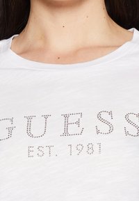 Bílý tričko s texturovaným, metalickým nápisem "GUESS" se studenými ozdobami a "EST. 1981" níže. Kolečkový výstřih, krátké rukávy.