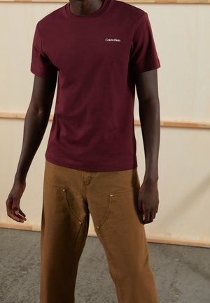 Muž v bordó tričku Calvin Klein a hnědých kalhotách stojí před světle dřevěnou stěnou a betonovou podlahou.