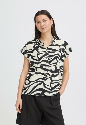 Woman wearing a short-sleeve black and white zebra print button-up shirt and black shorts, standing with one hand in pocket against white background.
