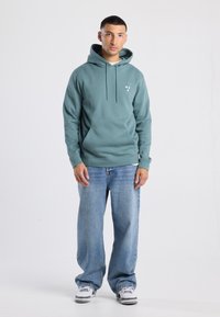 Teal hoodie met een voorzak en een capuchon met trekkoord, gecombineerd met lichtblauwe losse jeans en sneakers met grijze en witte accenten.