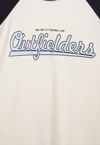 Tricou de baseball alb și negru cu text albastru „Outfielders” și „New York City Baseball Club” deasupra, pe piept.