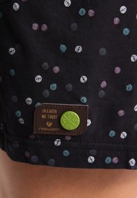 Svart tyg med flerfärgade prickar, med en brun etikett med text och en grön knapp, som visar en strukturerad, avslappnad design.