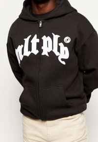 Black zip-up hoodie with a large white graphic text on the front, featuring kangaroo pockets and a drawstring hood.