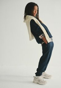 Chándal navy con una sudadera de punto crema texturizada caída sobre los hombros y zapatillas deportivas beige, exhibido sobre un fondo simple.