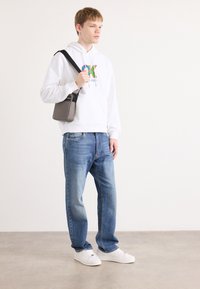 Haut à capuche blanc avec un logo multicolore, jeans bleu clair, sac à épaule gris et baskets blanches. Textures lisses et coupe décontractée.