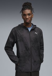 Jachetă neagră cu fermoar, cu un model texturat, dotată cu glugă și buzunare laterale. Logo-ul Puma pe piept în alb. Material neted, croi athletic.