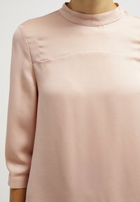 Jasnoróżowa bluzka z jedwabistego materiału, wyróżniająca się wysokim kołnierzem i rękawami o długości trzech ćwiartek, o gładkiej teksturze z subtelnymi detalami szycia.