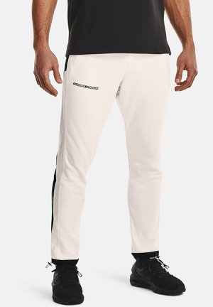 Mężczyzna w białych spodniach dresowych Under Armour z czarnymi pasami po bokach, czarnych trampkach i czarnej koszulce, stojący ze swobodnie opuszczonymi rękami wzdłuż ciała.