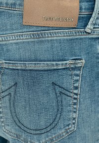 Niebieskie dżinsy typu denim z charakterystyczną skórzaną metką True Religion i zakrzywionymi szwami na tylnej kieszeni. Miękka tekstura i nowoczesny krój.