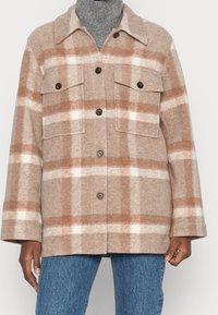 Chaqueta de lana de cuadros beige y blanca con botones y bolsillos de solapa en el pecho, llevada sobre un cuello de tortuga gris y jeans azules.