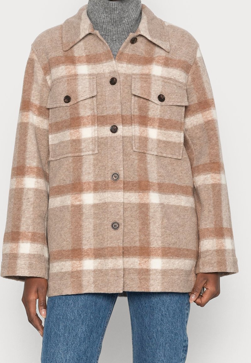Chaqueta de lana de cuadros beige y blanca con botones y bolsillos de solapa en el pecho, llevada sobre un cuello de tortuga gris y jeans azules.