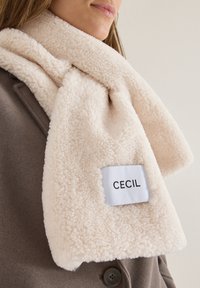 Cremefarvet, fluffy fleecehalstørklæde med et rektangulært hvidt mærke, der viser "CECI L." Båret over en brun frakke, som viser en blød tekstur.