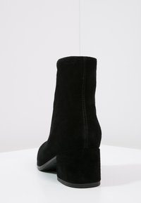 Bottine en daim noir avec talon bloc, couture arrière visible et semelle lisse, photographiée sur une surface blanche sur fond uni clair.
