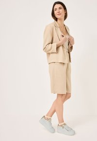 Blazer beige in lino abbinato a pantaloni corti su misura coordinati, completato da sneakers azzurre con dettagli dorati e calzini a fantasia.