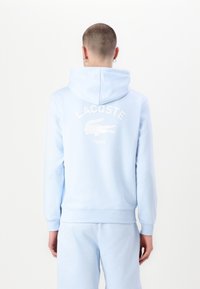 Šviesiai mėlynas hoodie su kengūros kišene, turi išaustą Lacoste logotipą su aligatoriumi ir "PARIS" tekstu nugaroje. Lygi tekstūra.