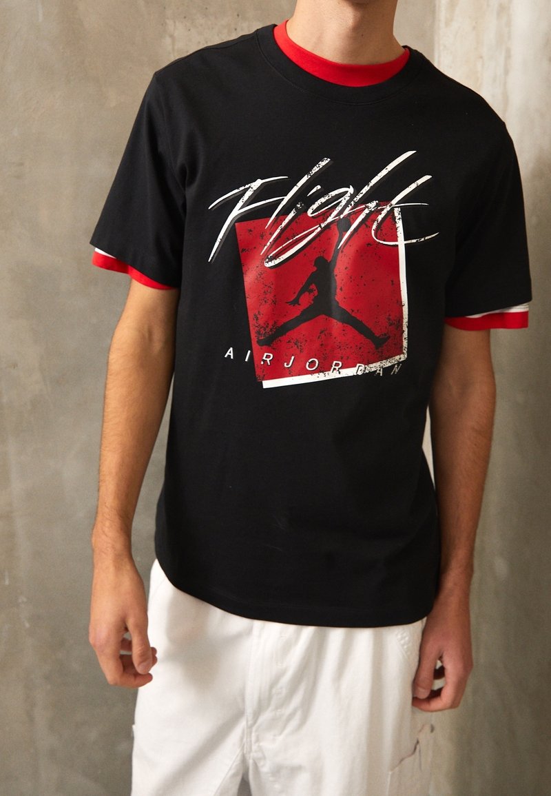 Personne portant un t-shirt noir "Flight Air Jordan" avec des manches superposées rouges et blanches et un pantalon blanc devant un mur gris uni.