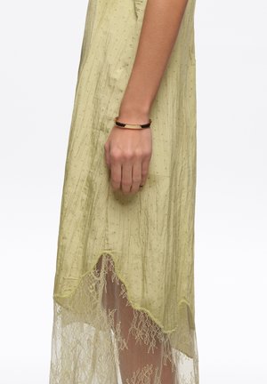 Ręka kobiety nosząca złotą bransoletkę obok żółtej sukienki z przezroczystą koronkową lamówką na białym tle.