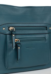 Borsa a tracolla in pelle verde acqua con due tasche con zip, dettagli in metallo dorato e una superficie liscia. Logo del marchio ben visibile sul davanti.