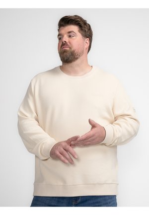Lichtbeige sweatshirt van een zachte stof met geribbelde manchetten en halslijn. Heeft subtiele branding op de borst en is gecombineerd met blauwe jeans.