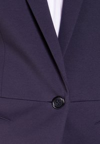 Blazer blu navy con tessuto strutturato, caratterizzato da un unico bottone nero e rever affilati. Cuciture visibili e cuciture sagomate.