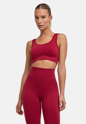 Mujer con sujetador deportivo rojo sin costuras a juego y mallas de talle alto, de pie sobre un fondo claro y liso.