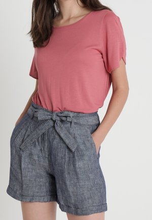 Vrouw draagt een roze top met korte mouwen en grijze paperbag-short met hoge taille, gestrikt met een stoffen riem, handen in de zakken.
