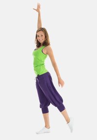 Green tank top with purple harem pants. The outfit features a fitted design with loose bottoms and white sneakers. Hands raised in a dynamic pose.