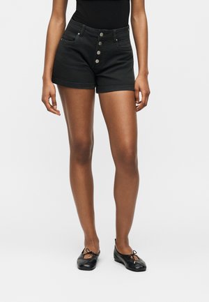 Nő fekete, magas derekú gombos rövidnadrágot, fekete felsőt és fekete balettcipőt visel, egyszerű világos háttér előtt áll.