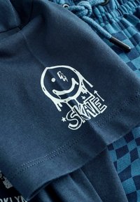 Tumši zila t-krekla ar lielu baltu grafiku, kas attēlo smaidīgu seju un vārdu "SKATE". Gluda auduma, īsām piedurknēm, ar redzamām šuvēm.