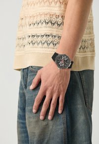 Montre noire avec un cadran octogonal, accents argentés et détails rouges ; portée au poignet d'une personne. Haut en tricot beige texturé, jeans bleus visibles.