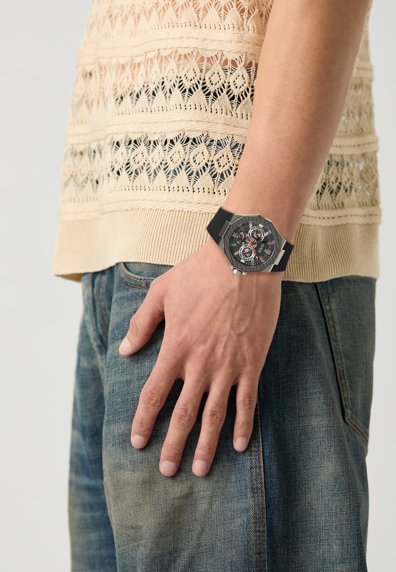 Montre noire avec un cadran octogonal, accents argentés et détails rouges ; portée au poignet d'une personne. Haut en tricot beige texturé, jeans bleus visibles.