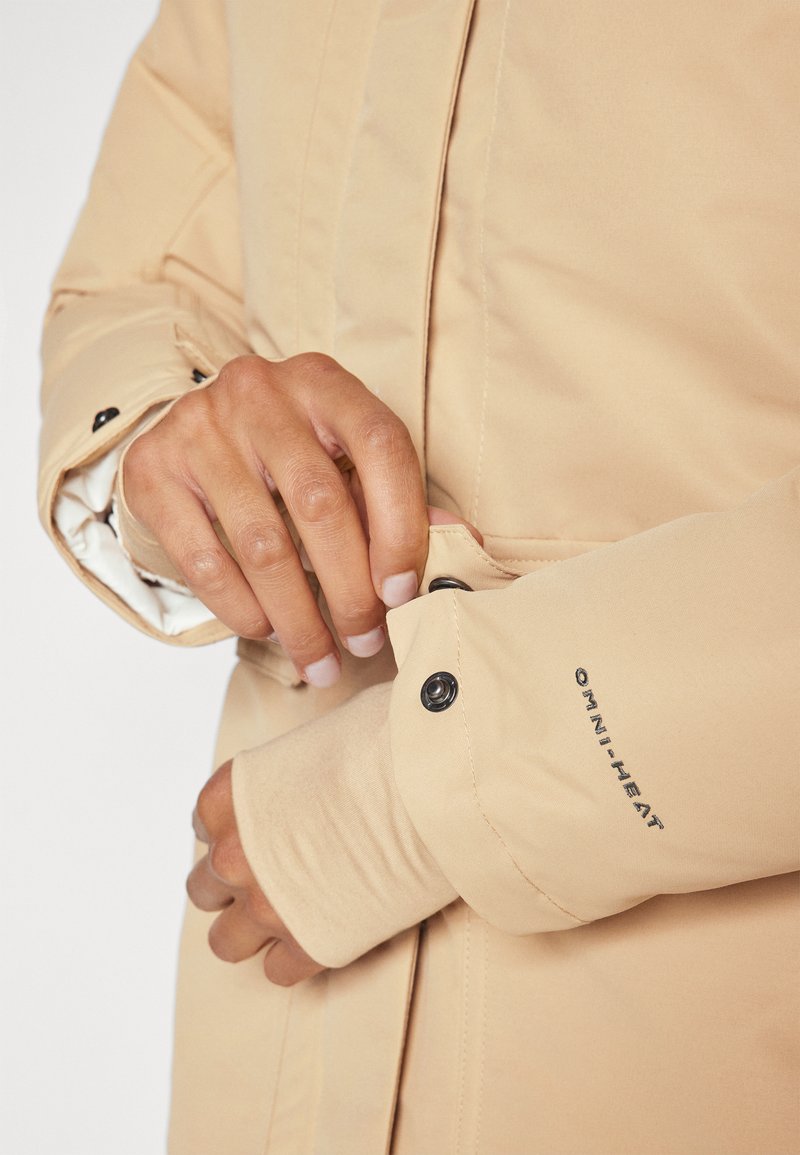 Veste isolée beige avec une finition lisse, des détails de poignet ajustés et des boutons-pression noirs. Comprend le marquage "OMNI-HEAT" sur la manche.