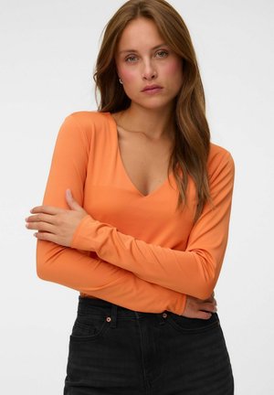 Kvinde med langt brunt hår iført en orange langærmet V-hals top og sorte jeans, står med armene over kors mod en ensfarvet baggrund.