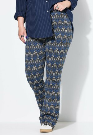 Persoon die een blauw-grijze broek met patroon draagt, een donkerblauw gestreept overhemd, witte sneakers en ringen aan de vingers.