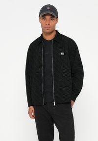 Juodas raštuotas užtrauktukas striukė su apykakle, derinama su juodu marškinėliu ir juodomis kelnėmis, užbaigia pilka kepurė su logotipu.