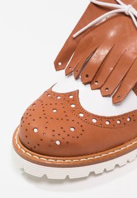 Chaussure brogue en cuir marron avec semelle blanche et détail de languette à franges nouée avec des lacets blancs sur fond blanc.