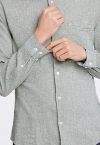 Camicia button-up verde chiaro, con texture, tasca sul petto, bottoni bianchi e maniche arrotolate, che mostra un design casual e un tessuto simile al lino.