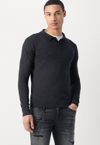 Mørkegrå langærmet polo shirt med krave og tre-knap lukning, sammen med slidte sorte jeans med kæde detaljer.