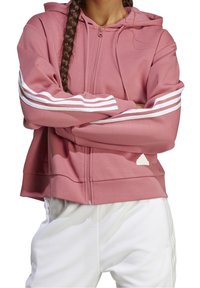 Femme portant un sweat à capuche rose zippé avec des bandes blanches sur les manches et un pantalon blanc, bras croisés sur la poitrine, tresse sur l'épaule.