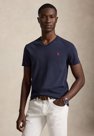 Homme portant un t-shirt bleu marine en V avec un logo rouge, pantalon blanc, ceinture marron et montre bleue, debout avec une main dans la poche.